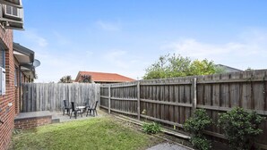 Ein Garten mit Holzzaun, einem Ziegelhaus, einer Terrasse mit Stühlen und einer kleinen Rasenfläche.