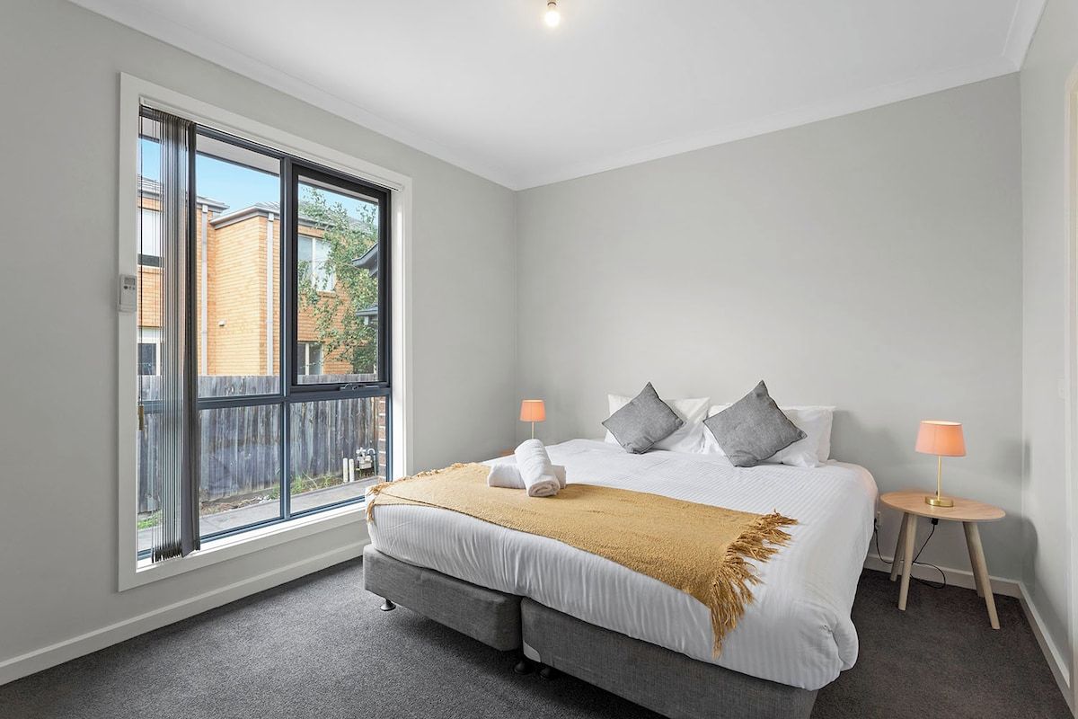 A bedroom with a large window, a bed with pillows, a bedside table, and a lamp.