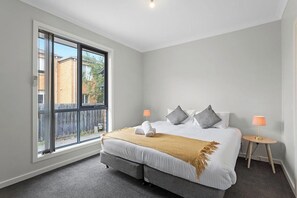 A bedroom with a large window, a bed with pillows, a bedside table, and a lamp.