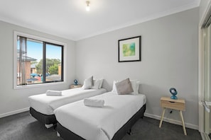 A hotel room with two beds, a window with a view, a bedside table, and a wall-mounted picture. - Domi Rentals - The Lantana Residence (Clayton)