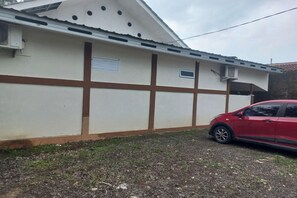 A building with a white facade and brown trim, a red car parked in front, and a gravel driveway. - Oyo 93749 Aksana Homestay Syariah (Purwokerto)