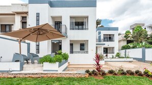A modern residential building with a flat roof, multiple balconies, and a paved courtyard with outdoor furniture and plants. - Nzeli Inn Hotel (Kigali)