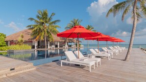 A poolside area with lounge chairs and red umbrellas, surrounded by palm trees and a view of the sea. - Noku Maldives (Noonu Atoll)