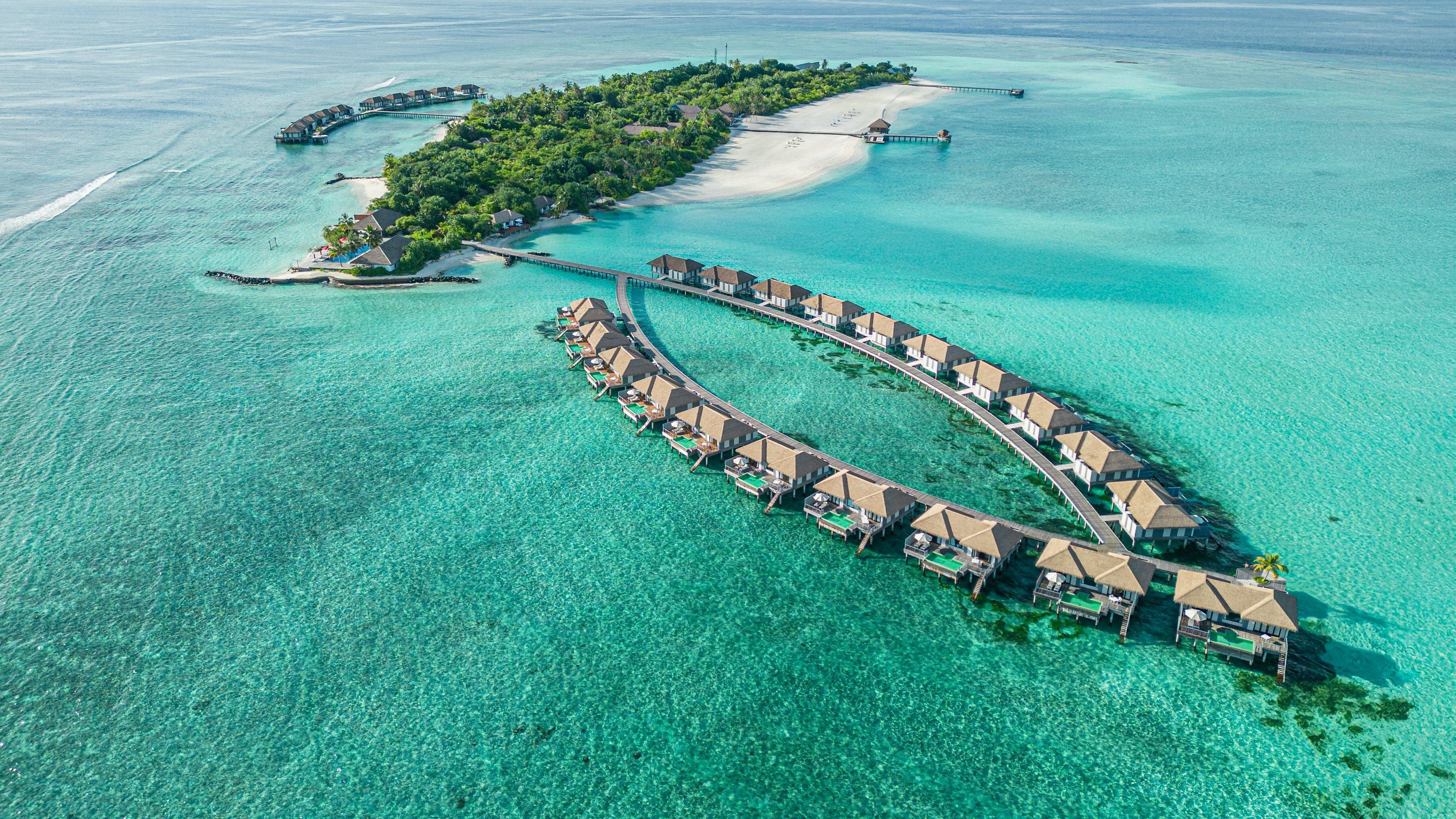 Un resort sobre el agua con una estructura de edificio en forma de media luna que se extiende hacia el mar turquesa, conectado por pasarelas.