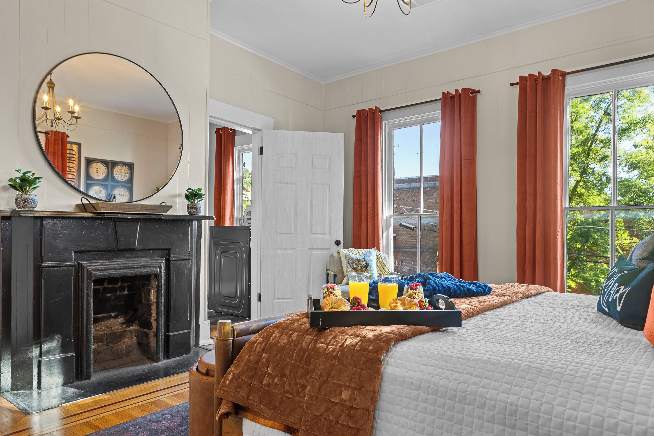 A bedroom with a fireplace, a bed with a brown blanket, a tray of breakfast, and a view of the outside.