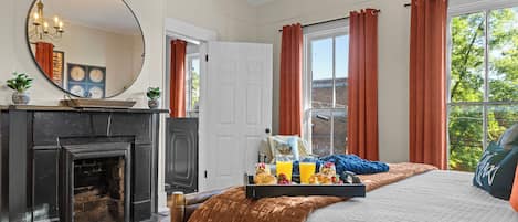 A bedroom with a fireplace, a bed with a brown blanket, a tray of breakfast, and a view of the outside.