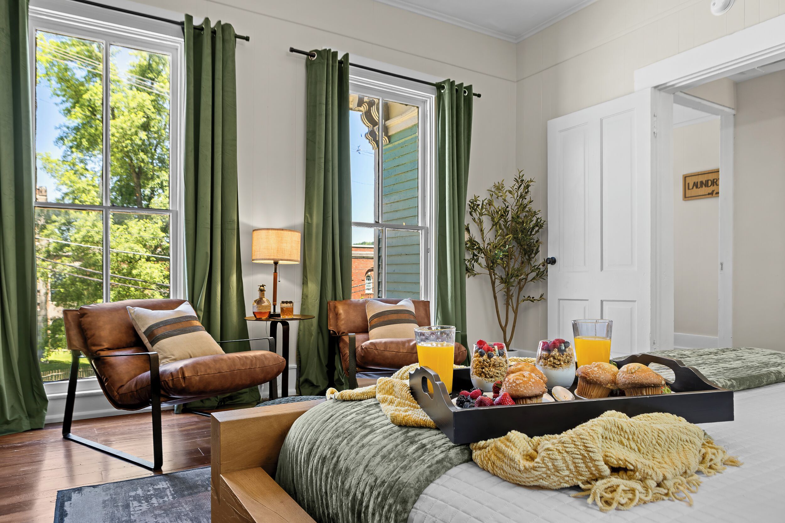 A hotel room with a bed, two leather chairs, a coffee table with breakfast items, and a view of the outdoors through large windows.