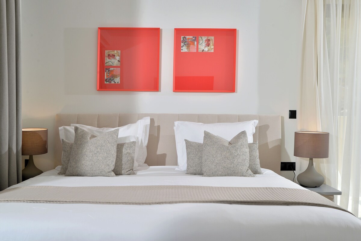 Een hotelkamer met een bed, twee bedlampen, een raam met gordijnen en twee ingelijste kunstwerken aan de muur.