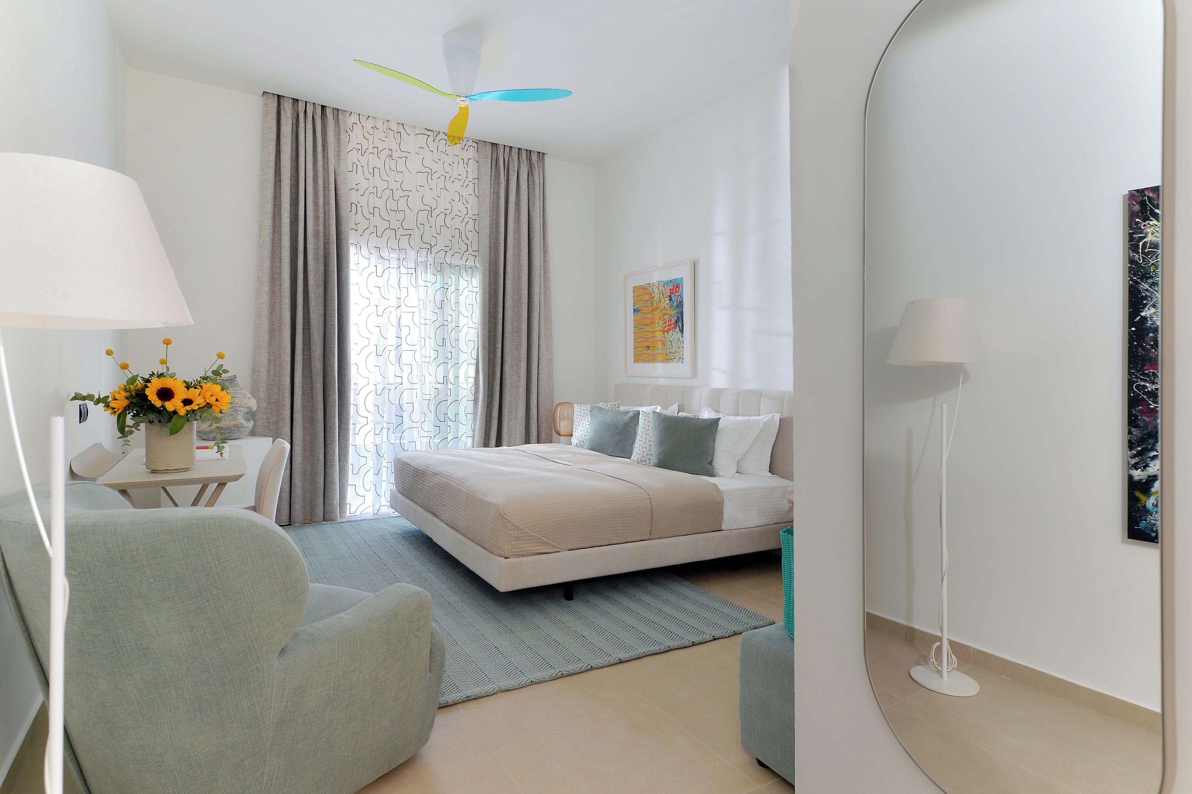 A modern hotel room with a bed, a desk with a lamp, a chair, a standing lamp, and a painting on the wall.