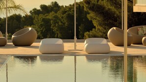 Un espace aménagé au bord de la piscine, avec des chaises longues et des parasols au design moderne, entouré d’arbres et surplombé d’un ciel dégagé.
