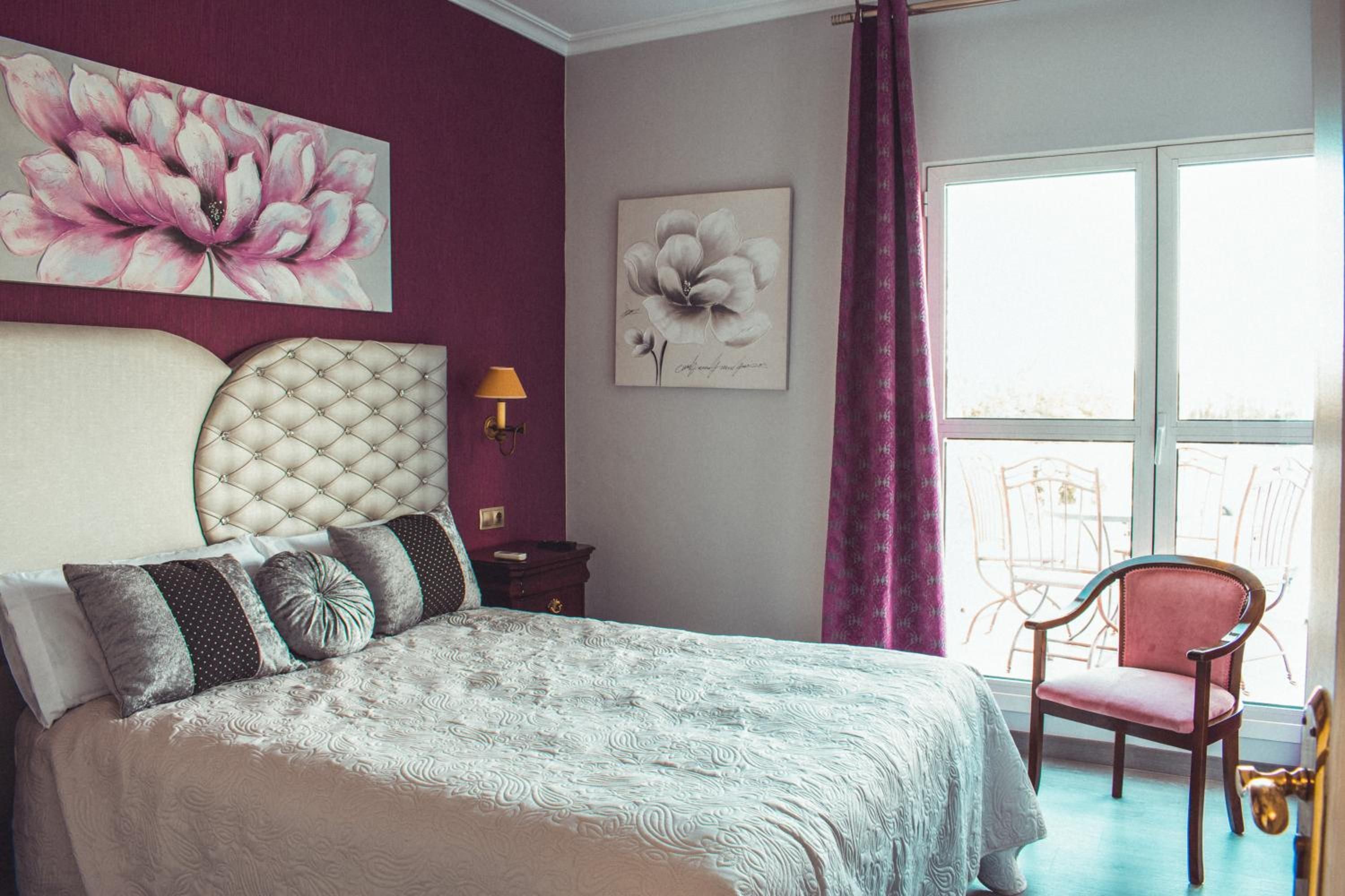 Un dormitorio con cabecera capitonada, una cama con ropa de cama estampada, una mesita de noche, una lámpara y una silla junto a la ventana.