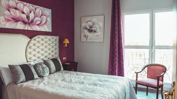 Un dormitorio con cabecera capitonada, una cama con ropa de cama estampada, una mesita de noche, una lámpara y una silla junto a la ventana.