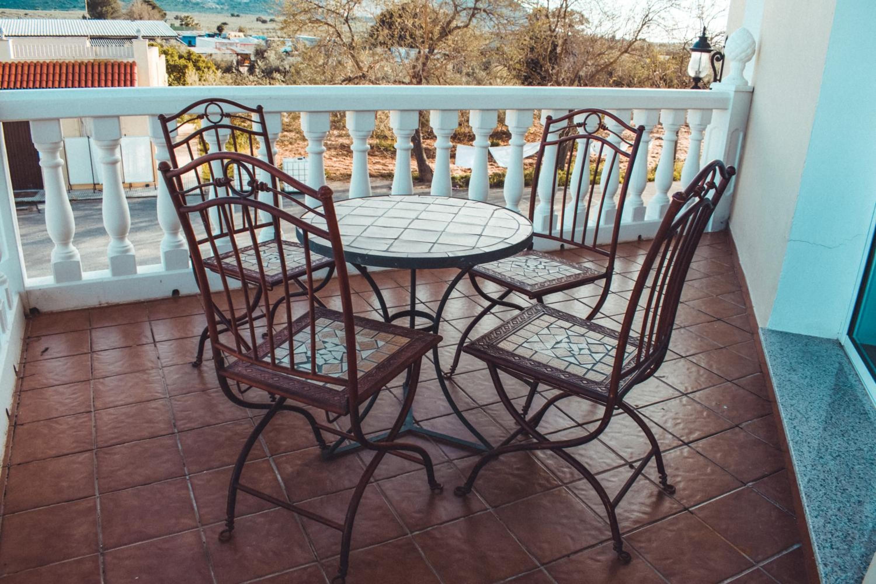 Un balcón con una mesa redonda y cuatro sillas, barandilla blanca y vista a una zona residencial.