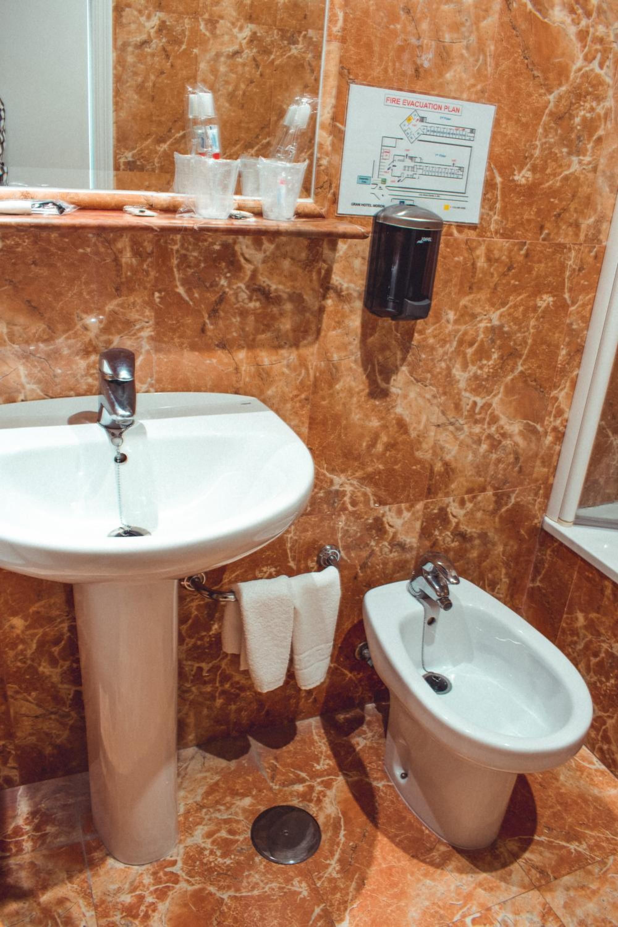 Un baño con un lavamanos blanco, un bidé y un plano de evacuación por fuego en la pared.