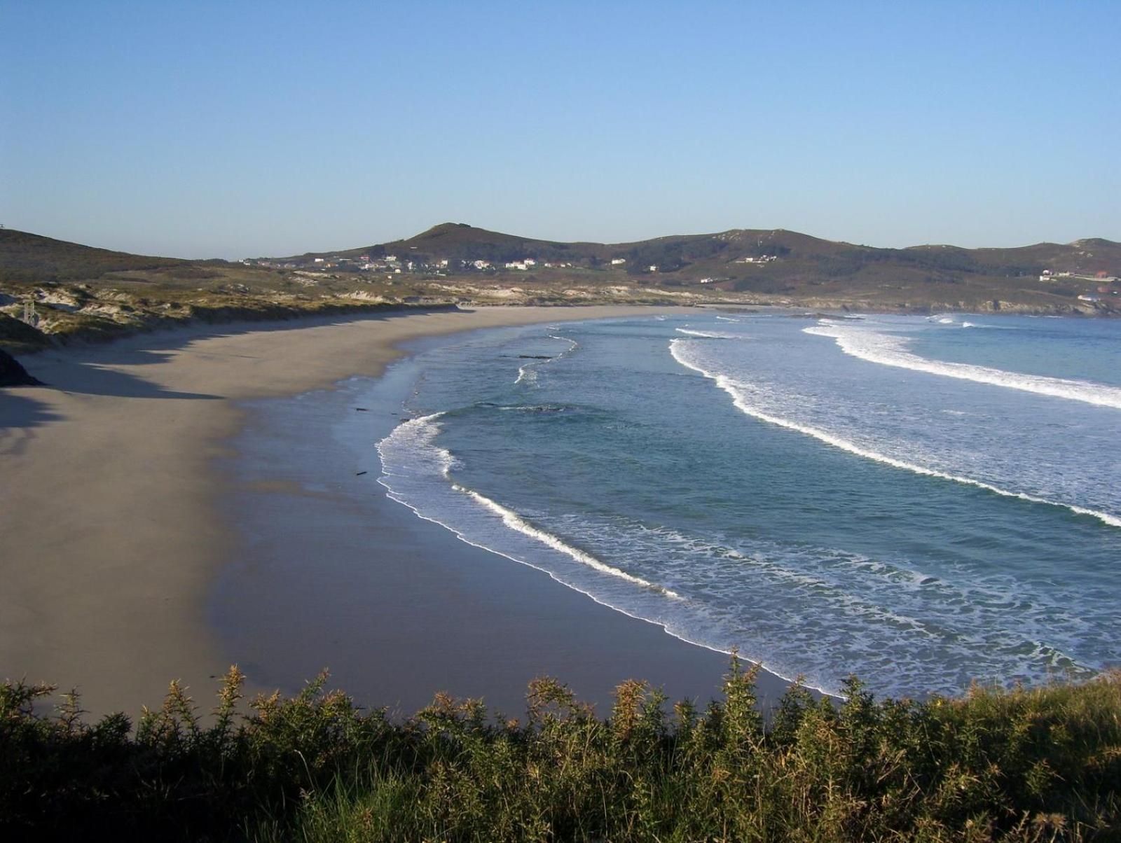 A coastal landscape with a sandy beach, waves, and distant hills.