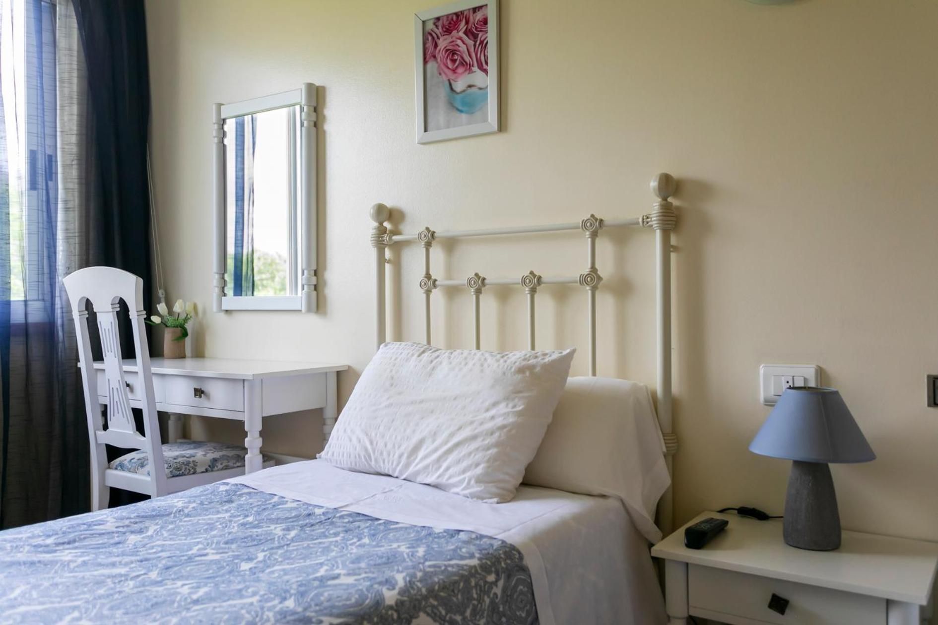 A bedroom with a bed, a desk, a chair, a lamp, and a painting on the wall.