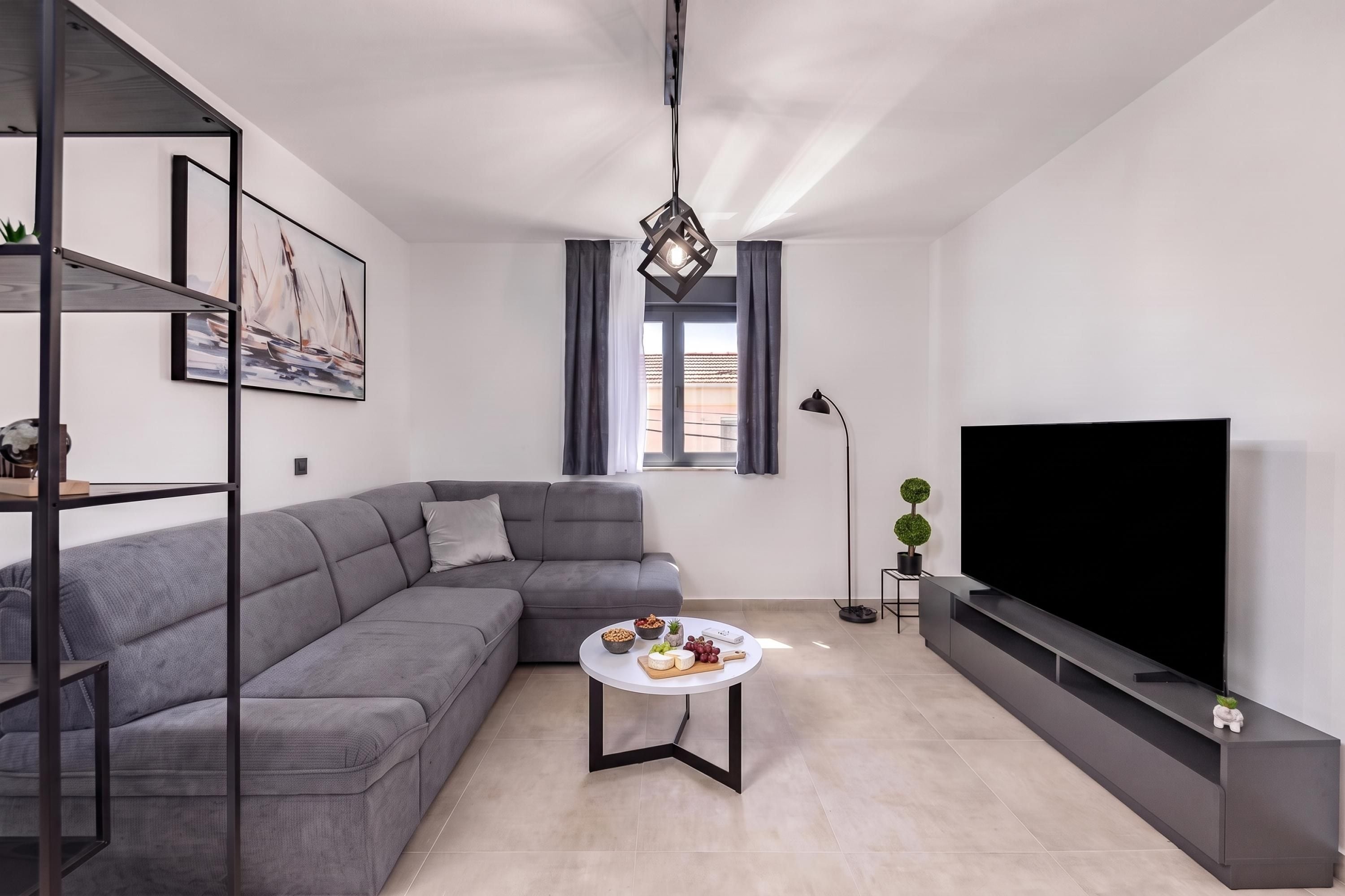 Uma sala de estar moderna com um sofá seccional cinza, uma mesa de centro redonda com petiscos, uma televisão de tela plana e uma luminária pendente.
