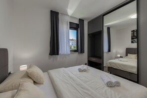 Ein Hotelzimmer mit Bett, Nachttischlampe, einem Fenster mit Vorhängen, einem Spiegel und Blick auf Gebäude.
