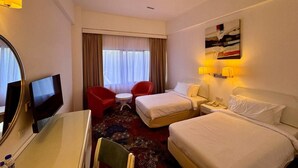 A hotel room with two beds, a desk with a flat-screen TV, a red armchair, a small round table, and a painting on the wall. - Royal Park Hotel (Kajang)