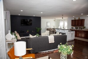 Ein modernes Wohnzimmer mit einem dunkelgrauen Sofa, einem Essbereich mit Tisch und Stühlen sowie einer Küche mit Holdekor-Schränken und einer weißen Arbeitsplatte.