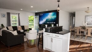 En moderne stue med en sofa, et spiseområde med et bord og stole, en køkkenø med skabe, og et stort fladskærms-tv, der viser 'Neo QLED 4K'.