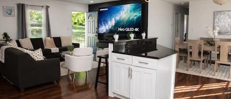 Una sala moderna con un sofá, un comedor con mesa y sillas, una isla de cocina con armarios y un televisor de pantalla plana grande que muestra 'Neo QLED 4K'.
