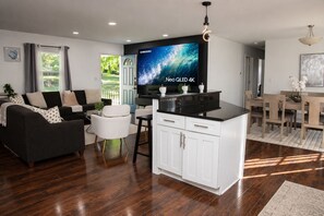 Ein modernes Wohnzimmer mit einer Couch, einem Essbereich mit Tisch und Stühlen, einer Kücheninsel mit Schränken und einem großen Flachbildfernseher, der 'Neo QLED 4K' anzeigt.