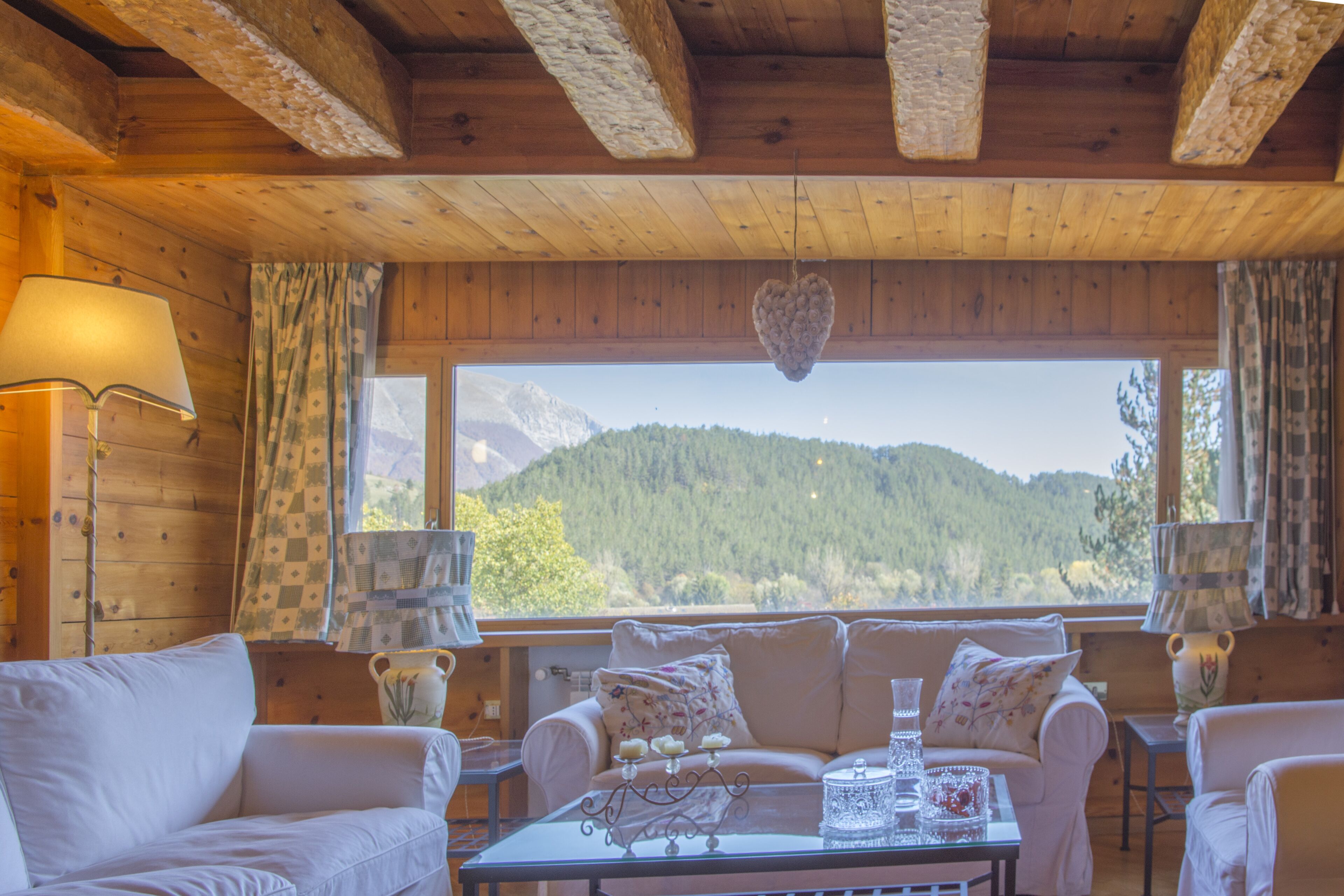 A wooden cabin interior with a sofa, a table with a vase and candles, and a view of a forested mountain.