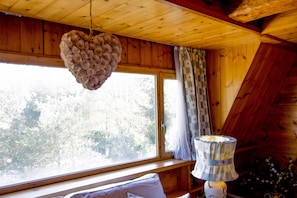 A cabin interior with a heart-shaped hanging decoration, a window with a view of trees, and a lamp on a wooden shelf. - Pescasseroli Appartamento Panoramico (Pescasseroli)