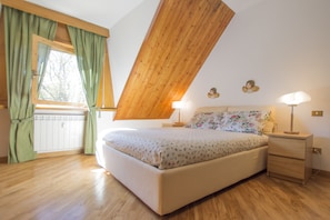 Ein Schlafzimmer mit schräg verlaufender Decke, einem Bett mit Blumenmuster auf der Bettwäsche, zwei Nachttischlampen und einem Fenster mit grünen Vorhängen.