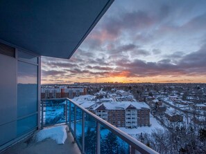 Varanda com guarda-corpo de vidro com vista para uma área residencial coberta de neve ao pôr do sol.