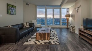 A modern living room with a sofa, coffee table, and TV, featuring large windows and a city view. - Icon Bay (Halifax)