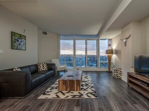 Sala de estar moderna com sofá, mesa de centro e televisão, com amplas janelas e vista para a cidade.