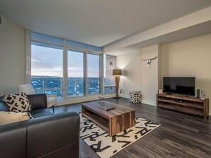 Uma sala de estar moderna com uma janela ampla, uma TV de tela plana, uma mesa de centro de madeira e um sofá.