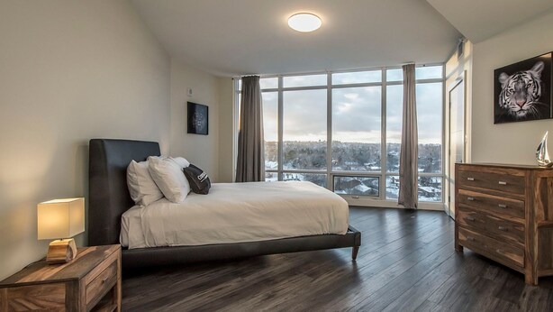 A bedroom with a large bed, a dresser, a nightstand, and a view of a snowy landscape through the windows. - Icon Bay (Halifax)