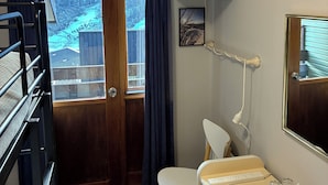 A hotel room with a wooden door, a television mounted on the wall, a desk with a chair, and a window with curtains.