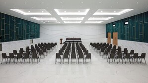 Konferenssihuone, jossa on riveissä järjestettyjä tuoleja, jotka on suunnattu puhujakoroketta kohti, marmorilattia ja upotetuilla valaisimilla varustettu katto.