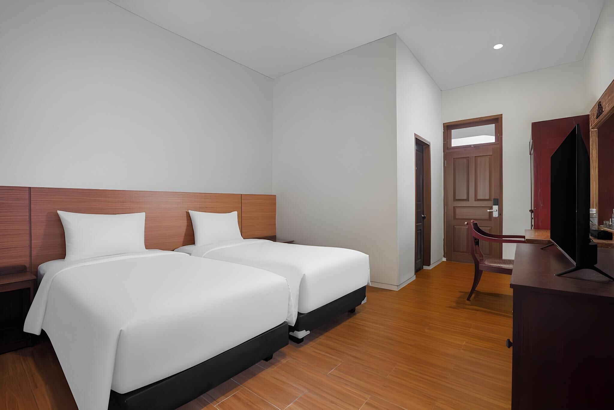 A hotel room with two beds, a wooden headboard, a desk with a chair, and a door leading to another room.