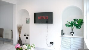 A modern living room with a mounted TV displaying 'NETFLIX,' a radiator, a chair, a vase with flowers, and a potted plant. - 3-bedroom House Bolton Town Centre Free Parking (Bolton)