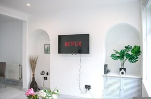 A modern living room with a mounted TV displaying 'NETFLIX,' a radiator, a chair, a vase with flowers, and a potted plant. - 3-bedroom House Bolton Town Centre Free Parking (Bolton)