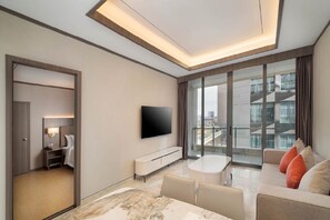 A modern hotel room with a flat-screen TV, a sofa, a dining table, and a balcony with a city view.