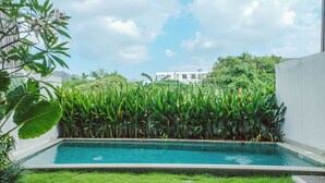 Una piscina rectangular con césped delantero, rodeada de abundante follaje y un edificio al fondo.