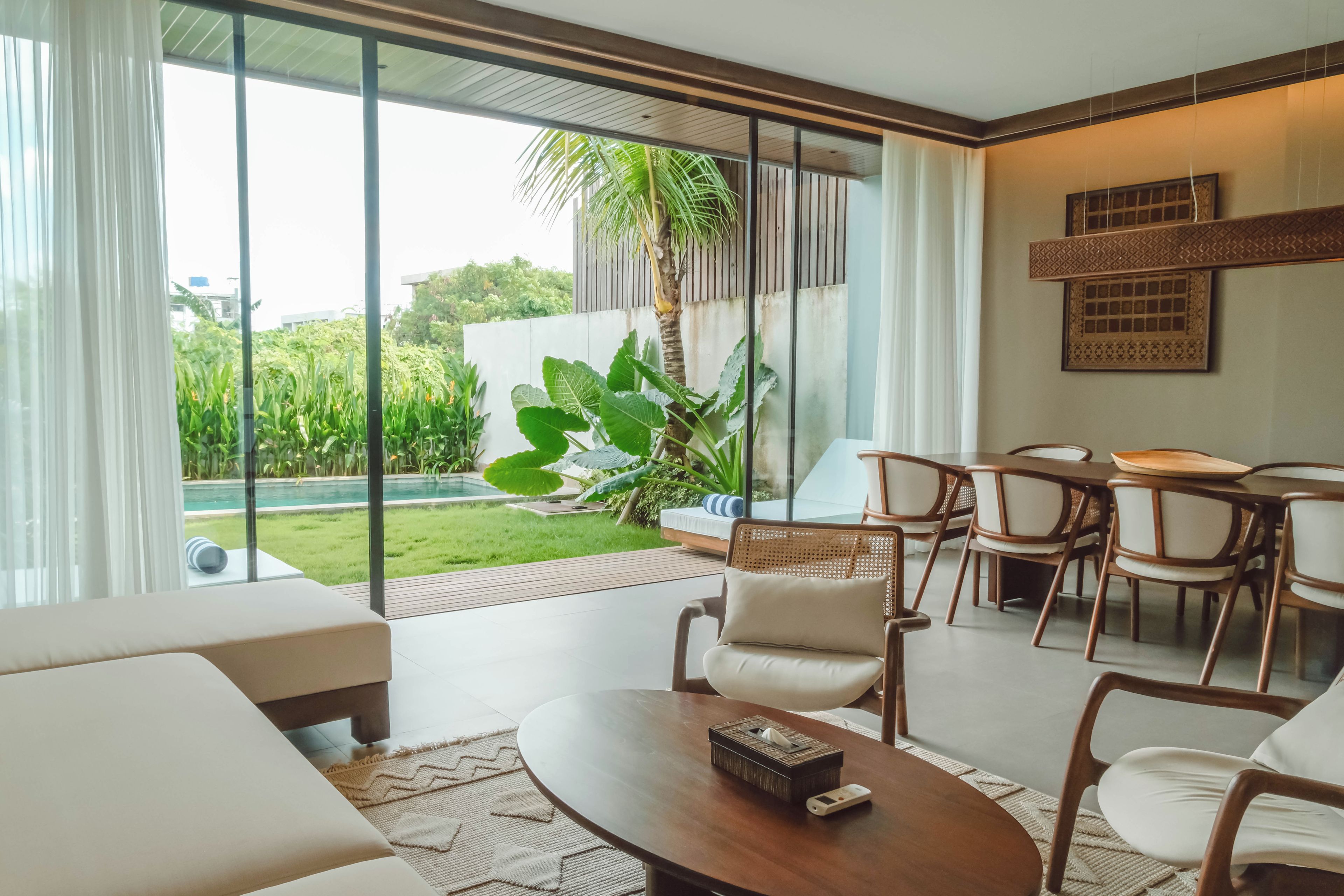 A modern living room with a large glass door leading to a pool, a dining area with chairs, and a large plant.