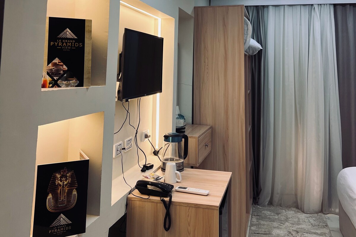 A modern hotel room with a flat-screen TV, a desk with a phone and a kettle, a bed with curtains, and a wall-mounted shelf with a framed picture.