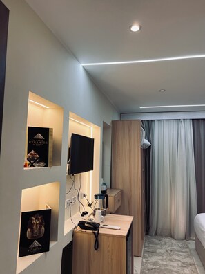 A modern hotel room with a flat-screen TV, a desk with a phone and a kettle, a bed with curtains, and a wall-mounted shelf with a framed picture. - Le Grand Pyramids View & Suites (Giza)
