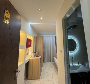 A hotel room with a wooden door, a minibar, a bed with curtains, a bathroom with a mirror and sink. - Le Grand Pyramids View & Suites (Giza)