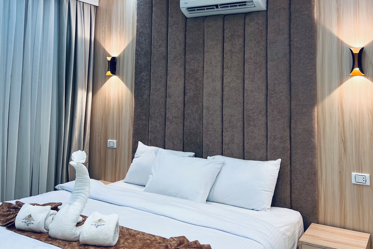 A modern hotel room with a bed, a nightstand, an air conditioning unit, and wall-mounted lighting.