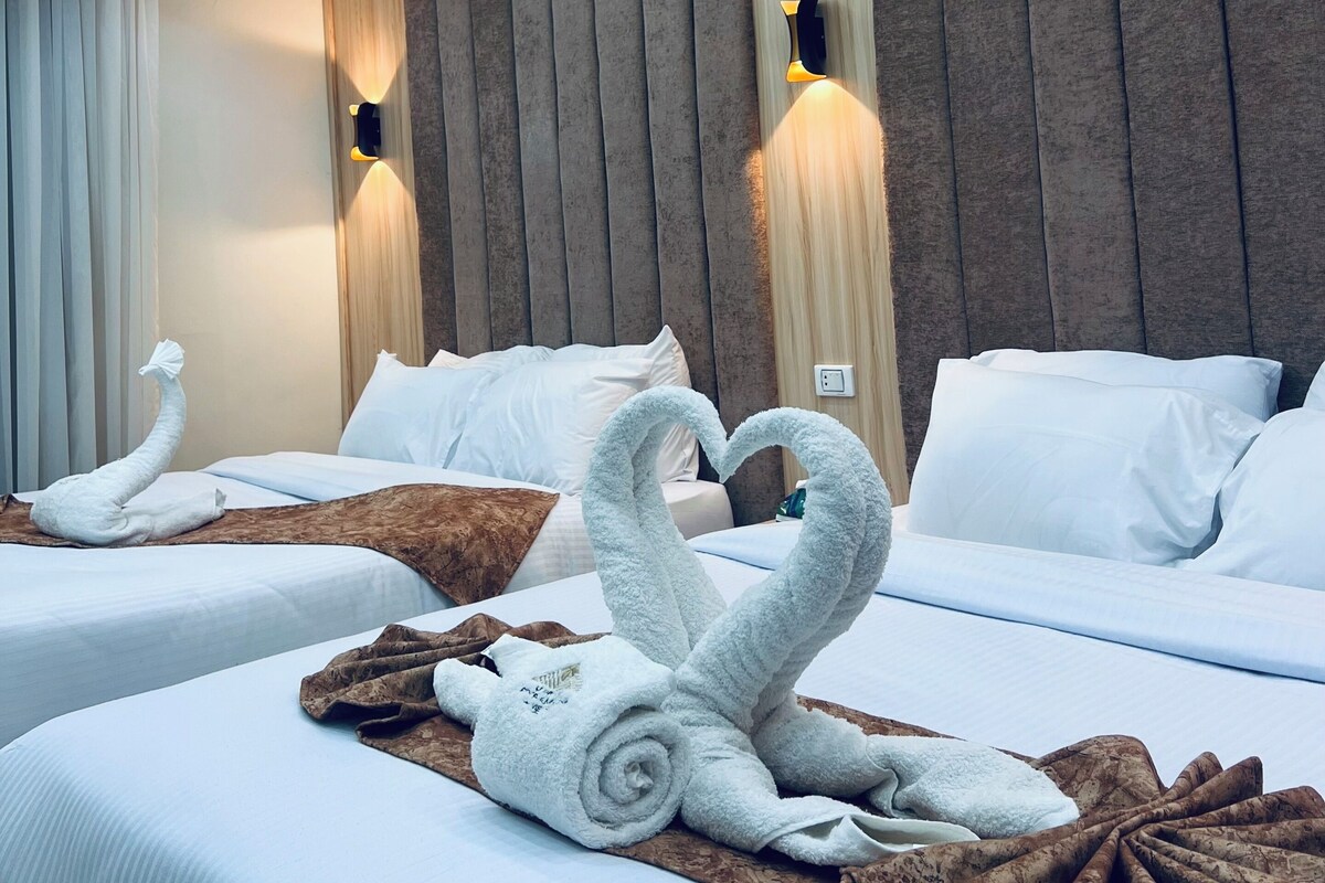 A hotel room with two beds, each with a swan towel art, a wooden headboard, and wall-mounted lighting.