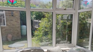 A conservatory with a sofa and a table, large windows with a view of greenery and a brick wall. - Spacious 4BR Stay in Morden Contractors and Group (Morden)