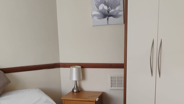 A bedroom with a bed, a nightstand, a lamp, a wardrobe, and a wall-mounted picture. - Spacious 4BR Stay in Morden Contractors and Group (Morden)